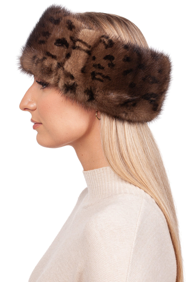 A woman with long blonde hair showcases a luxurious winter accessory: the Leopard Print Mink Fur Headband by FurbySD, made from ethically sourced mink fur and featuring subtle black spots. In profile and facing left, she pairs it with a beige sweater against a plain white backdrop.