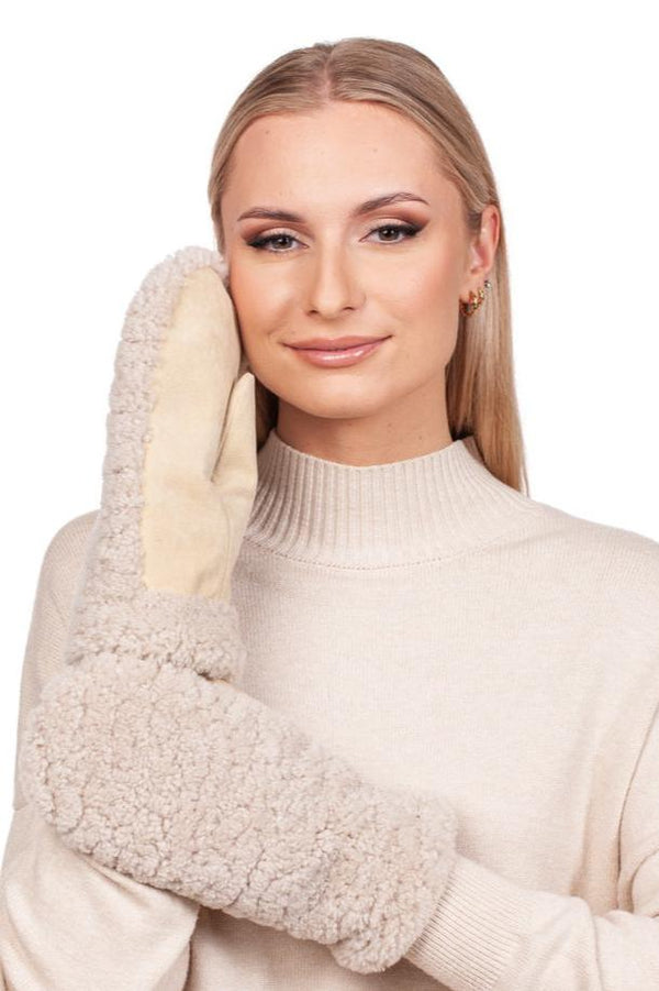 A woman with long blonde hair is wearing a cream sweater and holding a large, fluffy white mitten with a beige palm from the FurbySD Beige Curly Shearling Hat And Mittens Set against her cheek, smiling gently. Her cozy look is completed with winter accessories like the chic shearling hat included in the set.