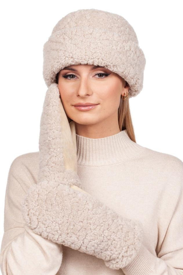 A woman in a cozy FurbySD ensemble, featuring the Beige Curly Shearling Hat And Mittens Set, exudes warmth. With one mitten softly pressed to her face against a plain white background, she embodies the essence of winter accessories.