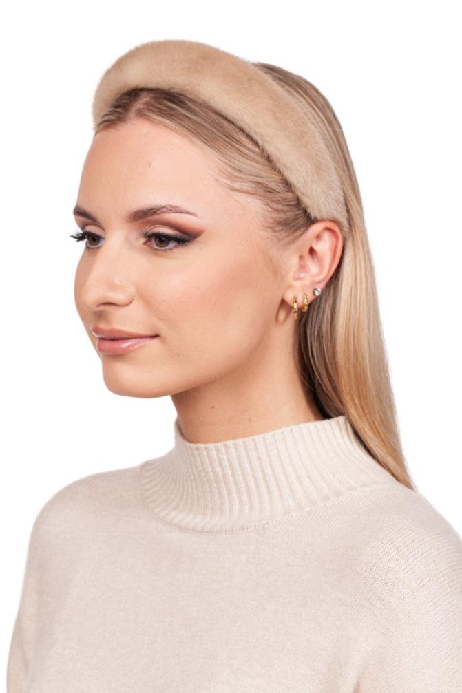 A person with long hair wears a FurbySD Beige Palomino Mink Fur Headband paired with a light-colored turtleneck sweater. They have multiple earrings and subtle makeup, looking to the side against a white background.