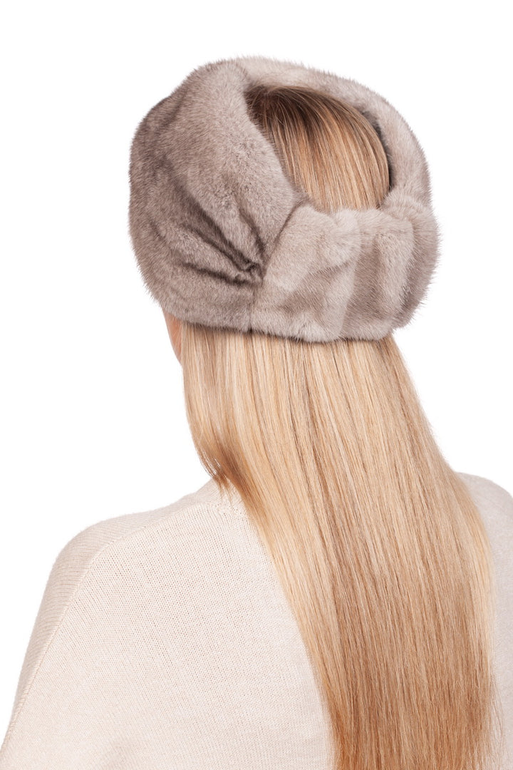 A person with long, straight blonde hair is wearing a Twisted Beige Mink Fur Headband from FurbySD, which wraps elegantly around their head. This luxurious winter accessory perfectly epitomizes sophistication as they face away, showcasing the headband's texture against a light-colored top.