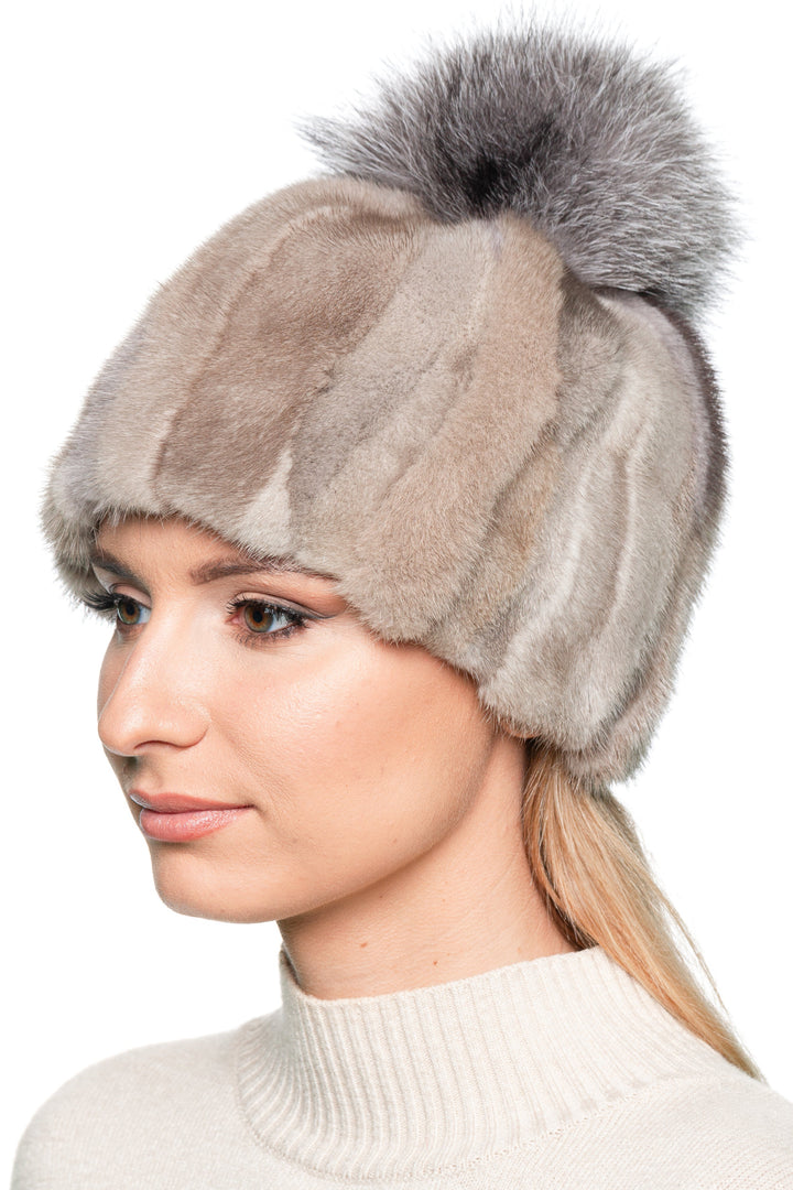 Side view of a woman wearing a beige patchwork mink fur beanie with a large silver fox pom pom. Fur panels in natural shades create a unique striped pattern.