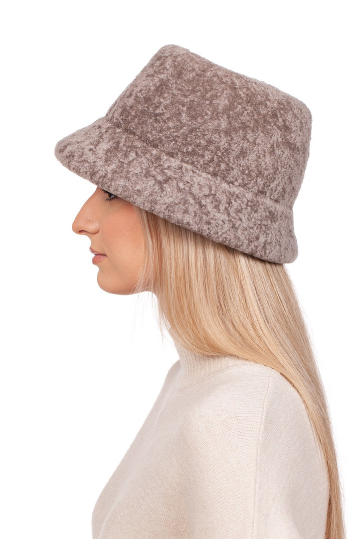 A person with long blonde hair is elegantly donning a luxurious accessory, the Beige Lambskin Fur Hat With Brim by FurbySD, along with a beige sweater, as they face left against a plain white background.