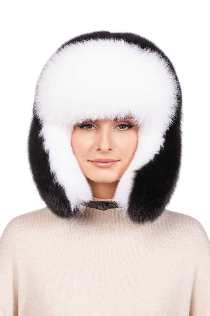 A person wearing the FurbySD Fox Fur Ushanka Trapper Hat in a classic combination of black and white, enveloping their head and ears. They are dressed in a light-colored sweater, smiling subtly against a plain white background—this hat is an undeniable winter essential.