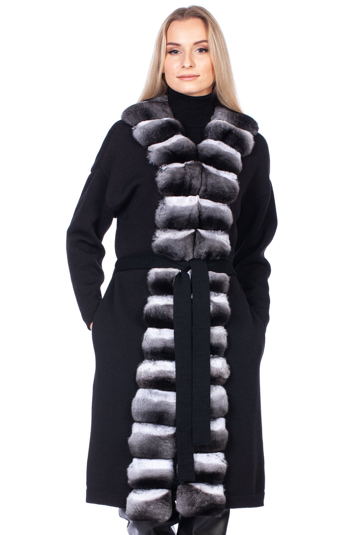A blond woman in the FurbySD Chinchilla Fur Trimmed Cashmere Coat, showcasing a black color against a white background. The coat features a luxurious chinchilla fur accent that runs vertically on the front. They complement the look with sleek black leather pants and shoes, with hands casually placed in the coat pockets.