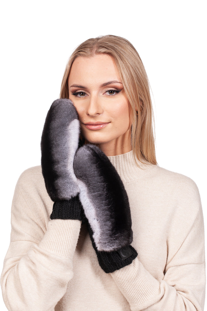 A woman with long blonde hair, dressed in a cream sweater, elegantly poses with luxurious FurbySD's Handknitted Black Merino Wool Mittens with Chinchilla Fur against her face. Her subtle makeup complements her gentle expression as she gazes at the camera, all set against a pristine white background.