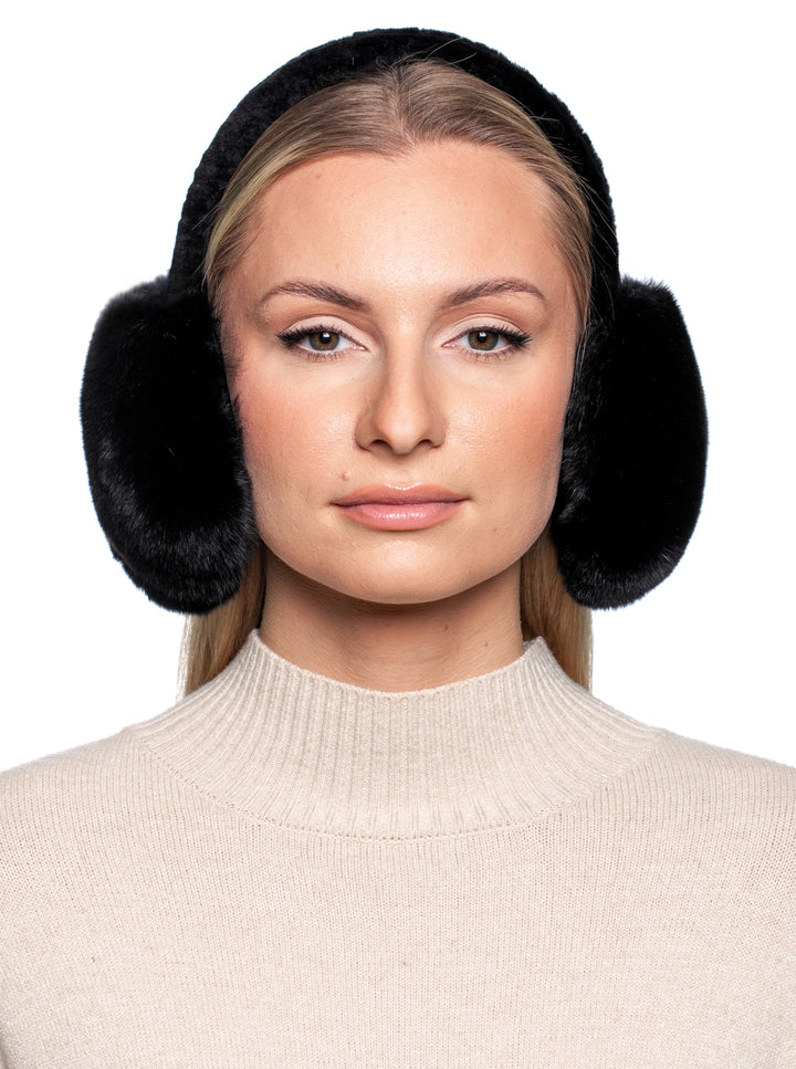 Against a white background, an individual dons the Black Chinchilla Fur Earmuffs with Astrakhan Band by FurbySD, paired with a beige sweater. They feature long, straight hair and maintain a neutral expression.