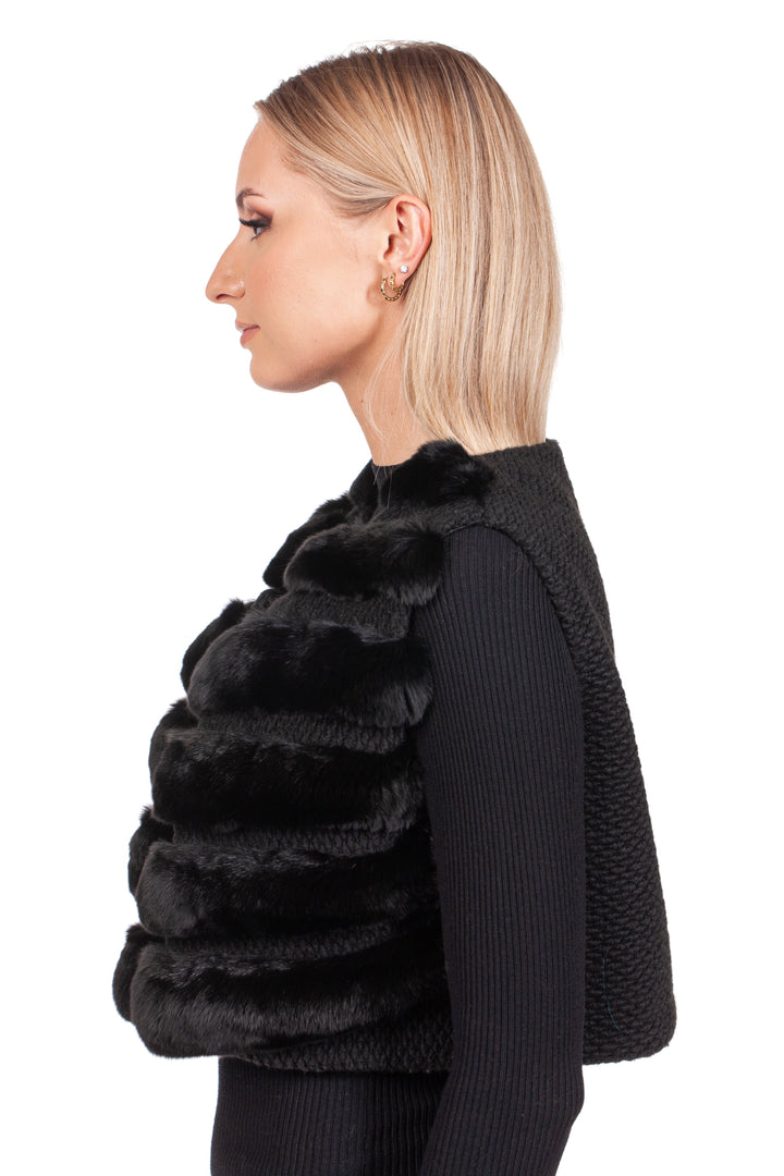 A woman with blonde hair is facing sideways, wearing the Black Chinchilla Fur Vest With Wool by FurbySD over a long-sleeved black top. The textured luxury fur gilet glistens elegantly against the stark white background.