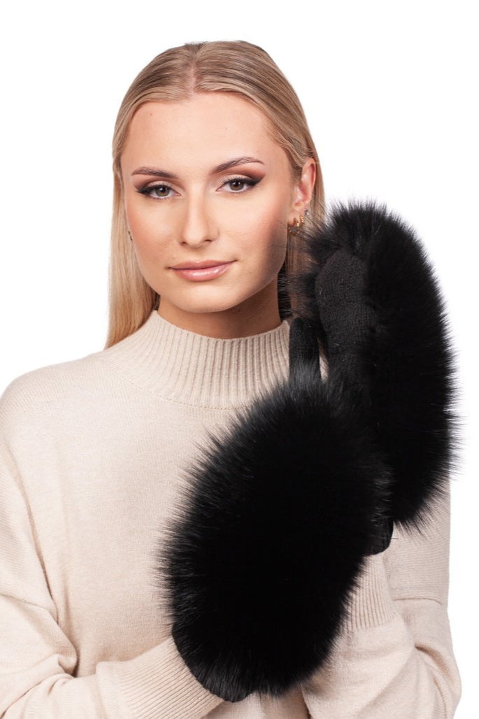 A woman with long blonde hair wearing a beige sweater holds up Black Fox Fur Flip Top Mittens by FurbySD. She smiles softly at the camera, reminiscent of an item from the SAGA auction house.