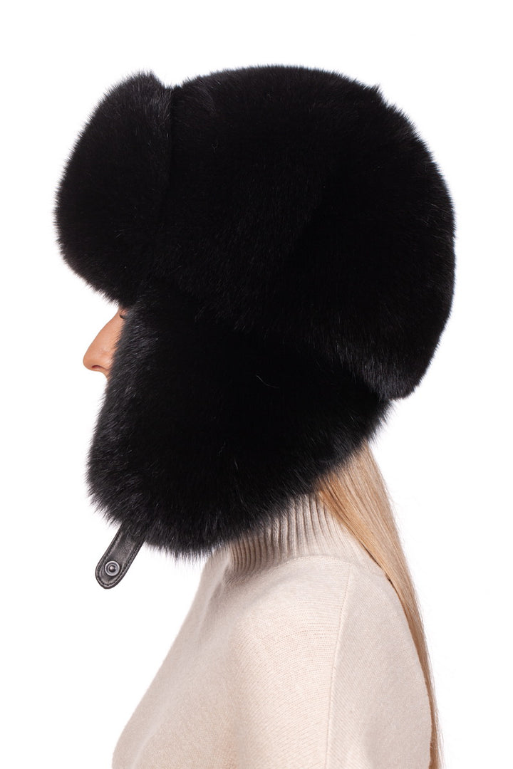 Wearing the Black Fox Fur Ushanka Trapper Hat by FurbySD, a person sports a large, fluffy headpiece with ear flaps that wrap snugly around their face. Positioned facing left in a light beige turtleneck sweater, this stylish winter accessory contrasts elegantly against the white backdrop.