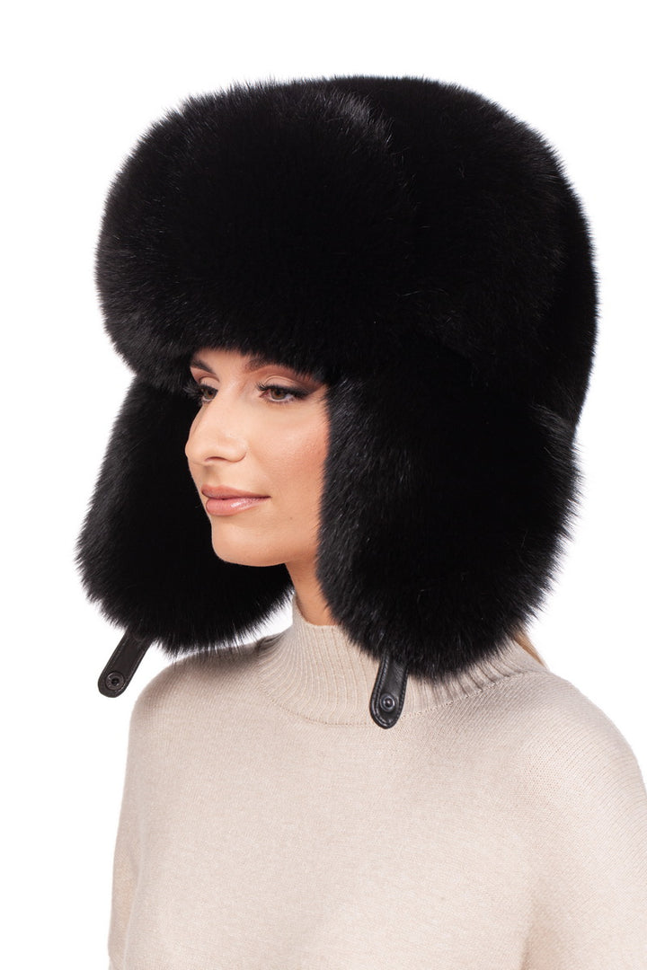 A person wearing the FurbySD Black Fox Fur Ushanka Trapper Hat with ear flaps stands against a white background. They are dressed in a light-colored sweater and looking to the side, showcasing their stylish winter accessory.