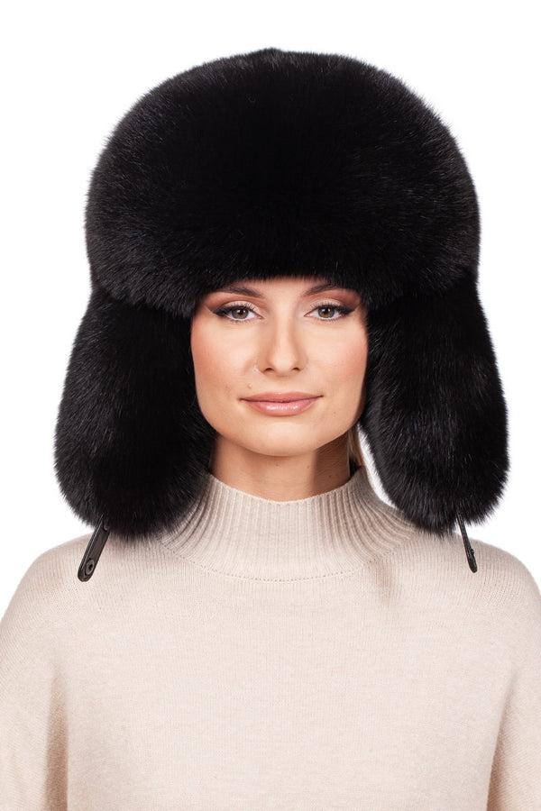 A person wears a Black Fox Fur Ushanka Trapper Hat by FurbySD, characterized by its large, fluffy design and ear flaps, paired with a light beige turtleneck sweater. The plain white background accentuates the texture and warmth of this winter accessory.