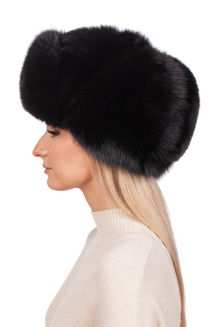 A profile view showcases an individual wearing a FurbySD Black Fox Fur Ushanka Trapper Hat paired with a light beige sweater. The minimalistic plain white background highlights the striking contrast between this chic winter accessory and their light attire.