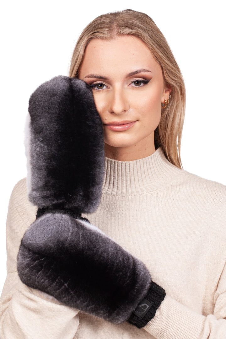 A person with long hair wearing a cream sweater poses against a plain white background, holding up FurbySD Handknitted Black Merino Wool Mittens with Chinchilla Fur to their face.