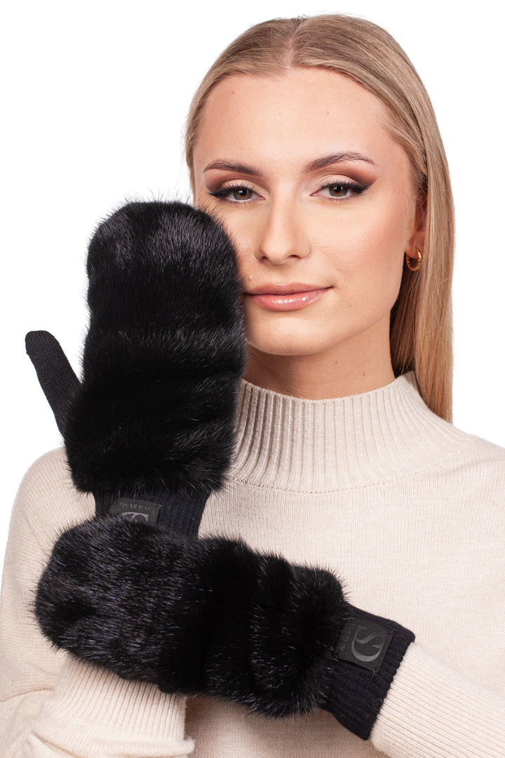 A woman wearing a cream merino wool turtleneck sweater is showcasing a pair of FurbySD Black Mink Fur Flip Top Mittens. Her long blonde hair gently cascades as she smiles softly at the camera, with a plain white background highlighting the cozy, chic vibe.