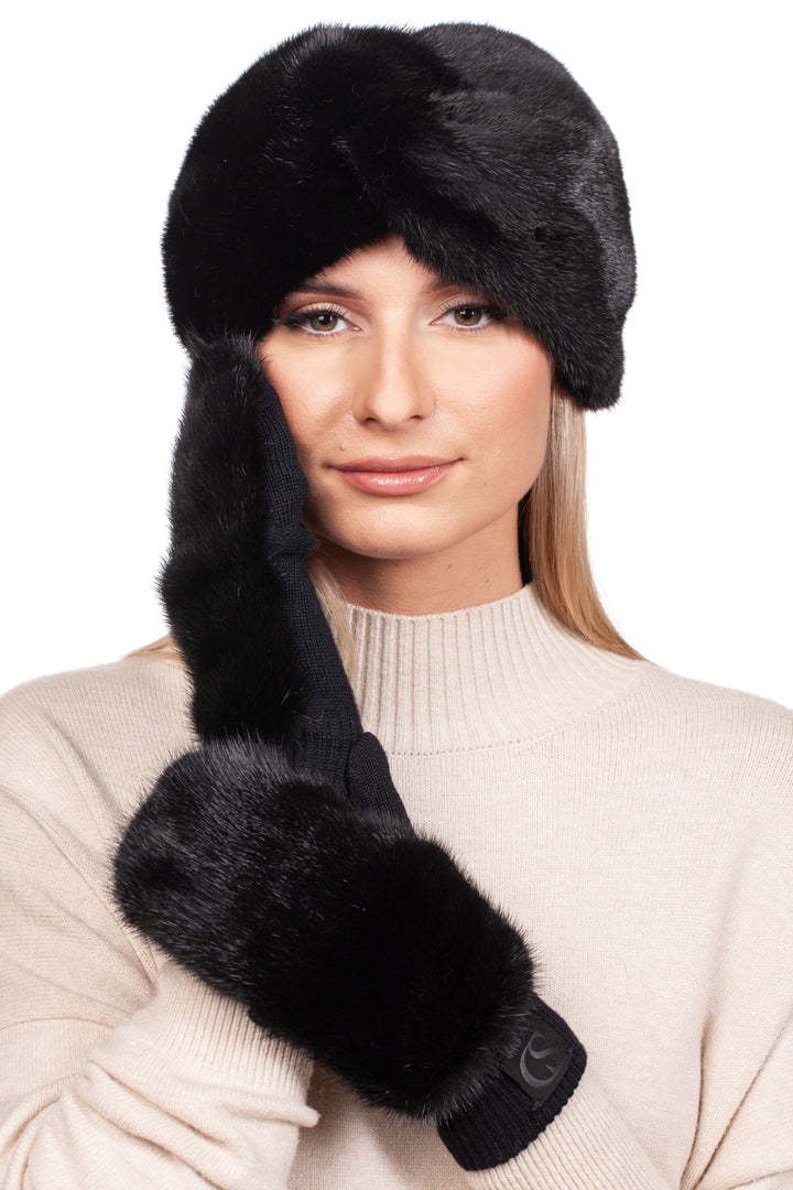 A woman exudes winter luxury, adorned in a black mink fur headband and mittens set from FurbySD, which perfectly complements her beige sweater. Her mittened hand gently touches her face as she offers a slight smile against the white backdrop.