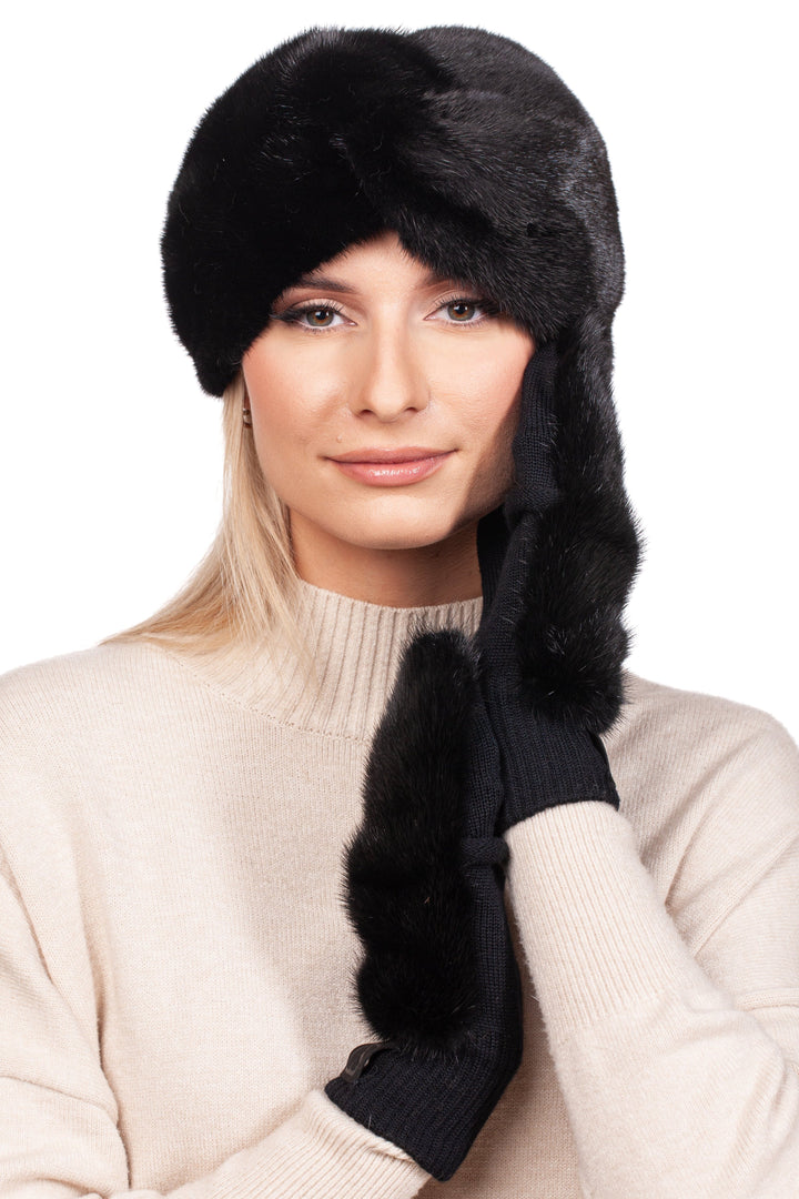 A person elegantly wearing the Black Mink Fur Headband and Mittens Set by FurbySD touches the side of their face. With long blonde hair cascading over a cream sweater, they stand against a simple white winter backdrop.