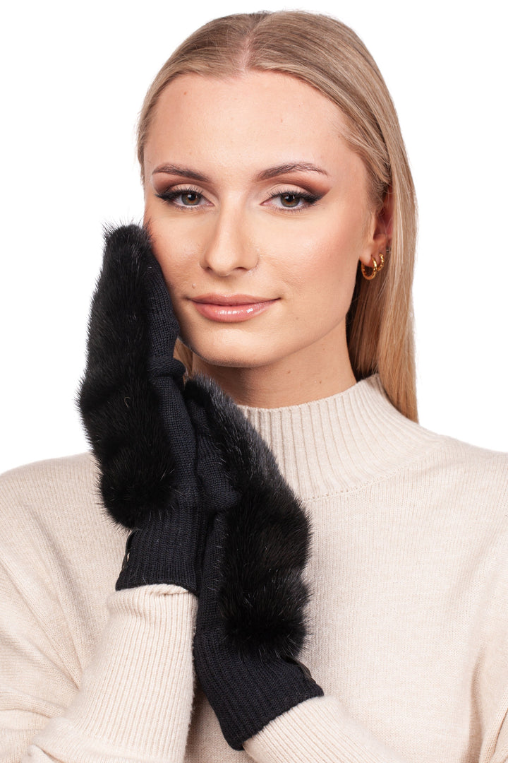 A woman with long blonde hair wears a handmade beige Merino wool sweater and Black Mink Fur Flip Top Mittens by FurbySD. She smiles gently with one gloved hand touching her cheek against a white background.