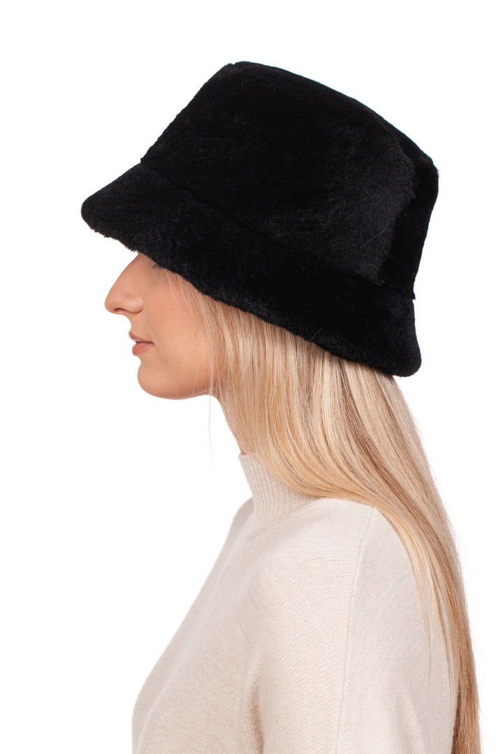 A person with long blonde hair is wearing the Black Mouton Lambskin Bucket Hat by FurbySD and a beige sweater, facing left against a plain white background.