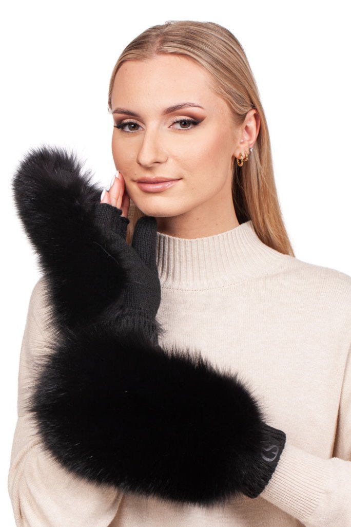 A woman with long blonde hair is wearing a beige turtleneck sweater and large Black Fox Fur Flip Top Mittens by FurbySD. She is touching her face with one hand and smiling slightly, set against a plain white background.