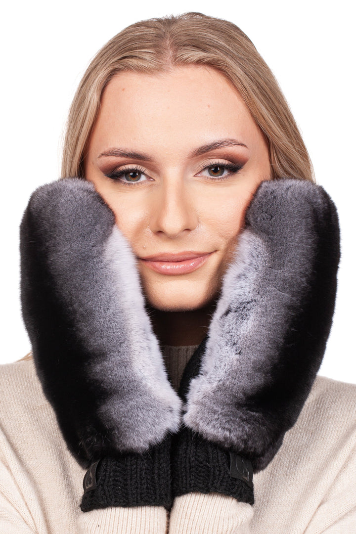 A woman with long blonde hair is smiling, holding FurbySD's Handknitted Black Merino Wool Mittens with Chinchilla Fur against her cheeks. She is wearing a cream-colored sweater against a white background.