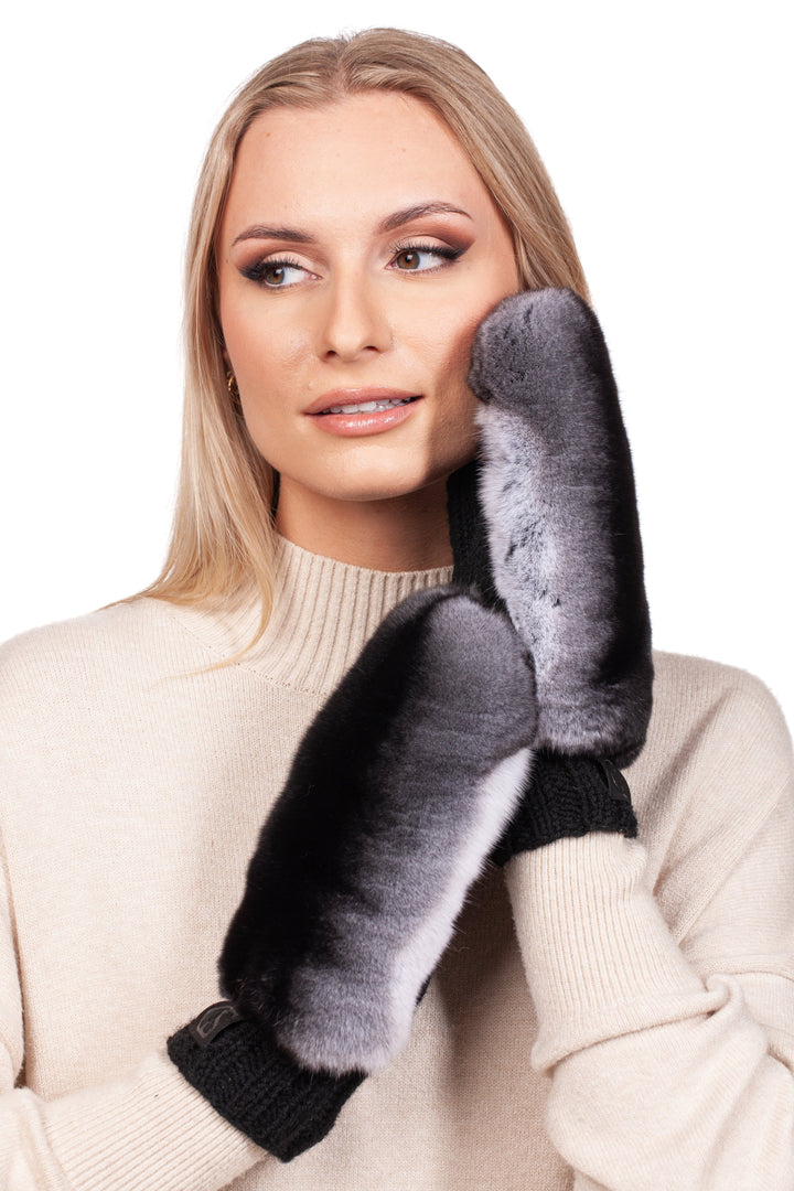 A woman with long blonde hair is wearing a cream-colored sweater and FurbySD's handknitted black merino wool mittens adorned with chinchilla fur, gazing thoughtfully to the side against a white background.