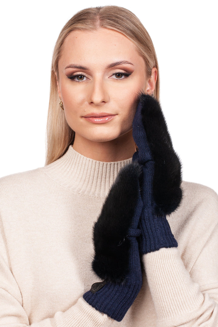 A person with long blonde hair is wearing a cream sweater and handcrafted luxury mittens from FurbySD, featuring a dark blue hue with elegant black mink fur trim. The mittens gracefully touch their cheek as they look directly at the camera.