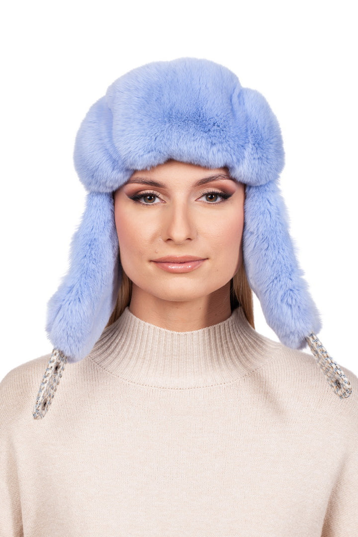 A person is wearing the Blue Chinchilla Fur Ushanka Hat from FurbySD, featuring a light blue color with fluffy ear flaps and ribbon ties. They have long hair and are dressed in a light beige turtleneck sweater, perfectly capturing the essence of winter headgear against a plain white background.