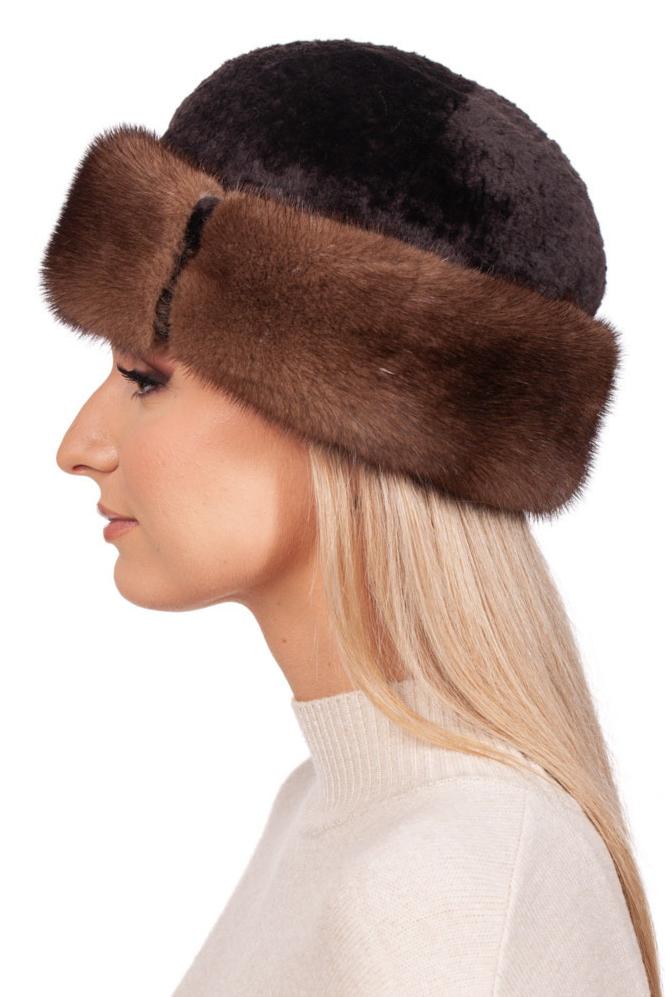 A woman with long blonde hair models a FurbySD Brown Mink and Astrakhan Fur Roller Hat, featuring a wide brown fur trim. She is turned to the side in a light beige sweater, perfectly embodying premium winter accessories against a plain white background.