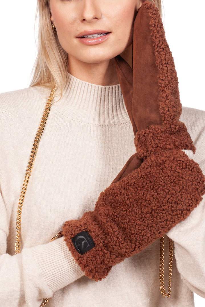 A person wearing a cream sweater is holding the Brown Shearling Fur Mittens With Detachable Chain by FurbySD to their ear, as if it were a phone receiver. Their blonde hair is complemented by gold earrings and the unique handmade quality of these mittens adds an artisanal touch to their ensemble.