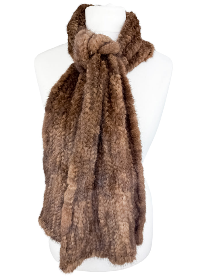 A brown knitted mink fur scarf by FurbySD is elegantly draped around a white mannequin. The scarf is long, fluffy, and appears very soft, showcasing its rich texture and ethically sourced craftsmanship.