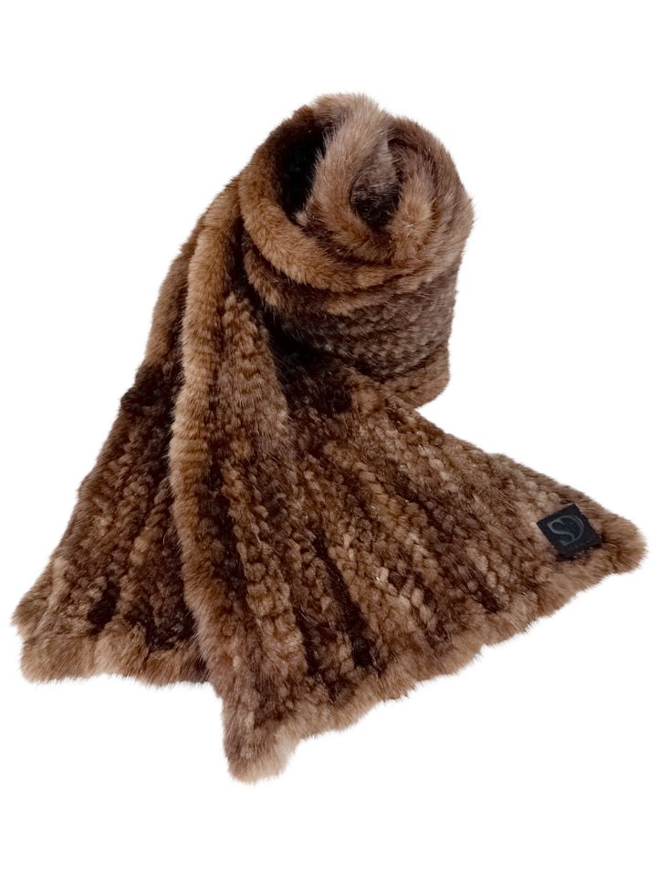 A Brown Knitted Mink Fur Scarf from FurbySD is elegantly draped, highlighting its ribbed texture and featuring a subtle black tag on one end. Ethically sourced, it boasts rich shades of brown that exude a warm and cozy look.