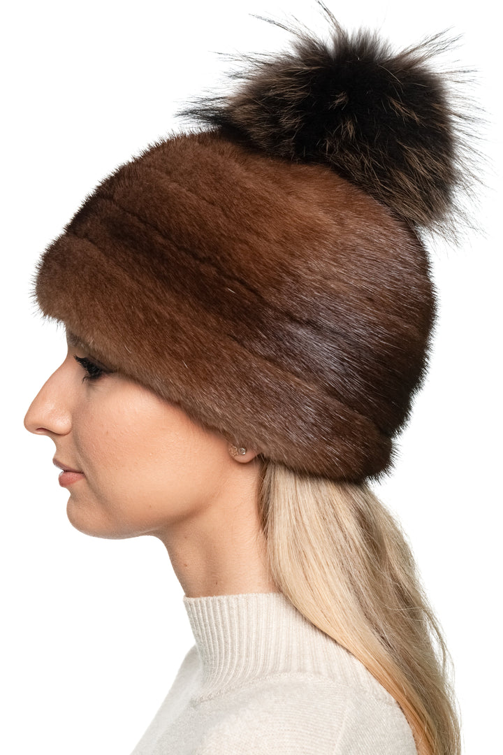 Brown mink fur hat with a fox fur pom pom worn by a person on a white background.