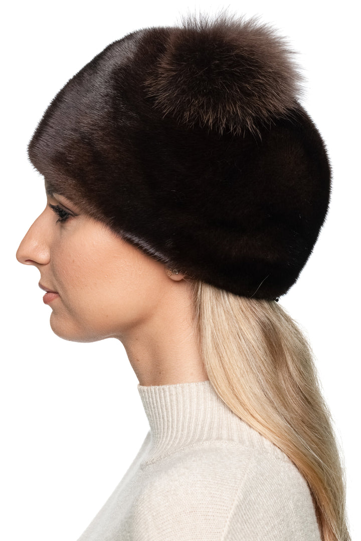 Woman wearing a dark mahogany mink fur hat with matching two fox fur pom poms on a white background