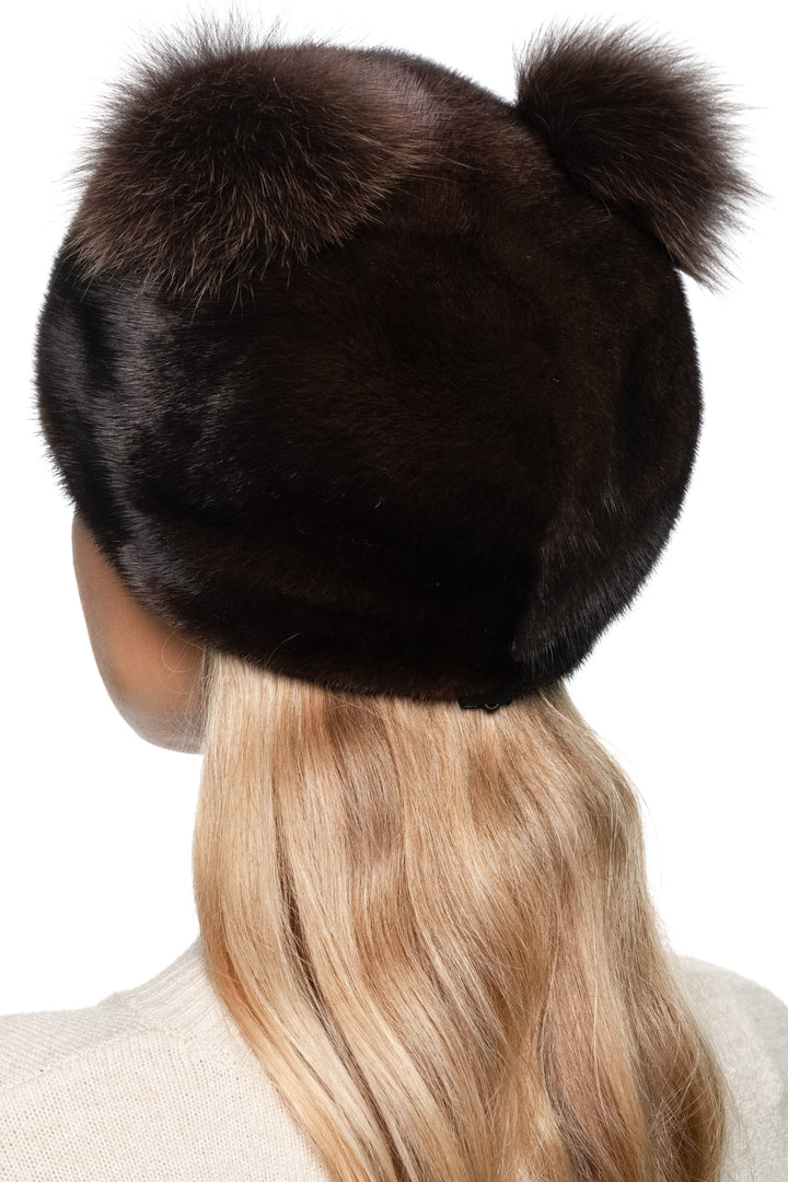 Woman wearing a dark mahogany mink fur hat with matching two fox fur pom poms on a white background.