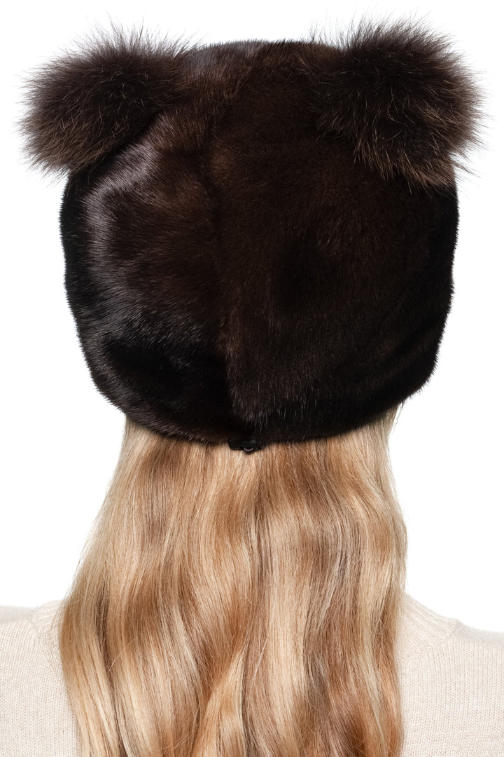 Woman wearing a dark mahogany mink fur hat with matching two fox fur pom poms on a white background.