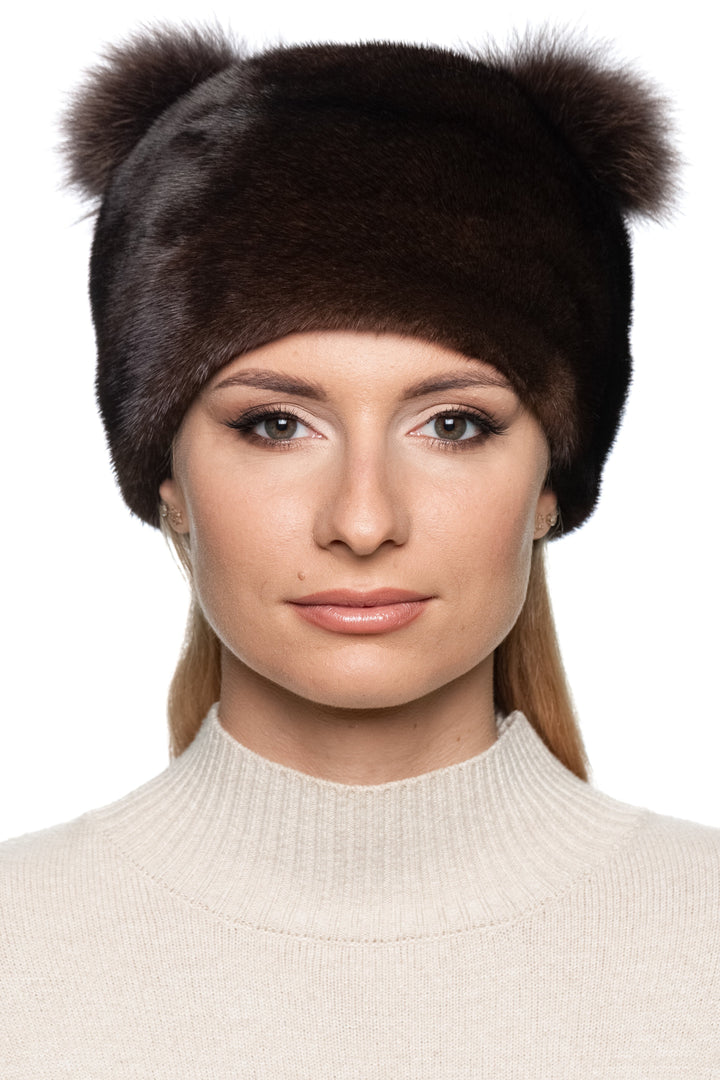 Woman wearing a brown mink fur hat with matching fox fur pom poms on a white background.