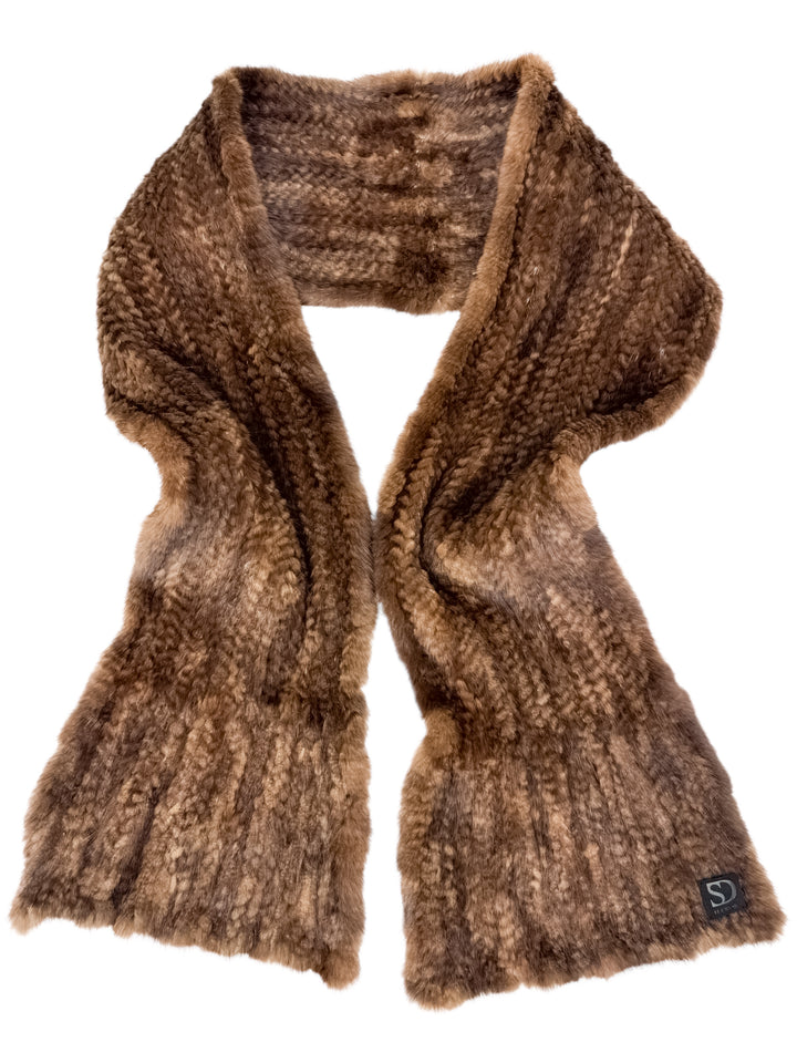 The Brown Knitted Mink Fur Scarf by FurbySD features a long and thick design with a textured pattern for an elegant look. Its soft and fuzzy appearance is enhanced by the delicate folding at both ends. A small black label displaying the initials "SD" can be seen at one end.