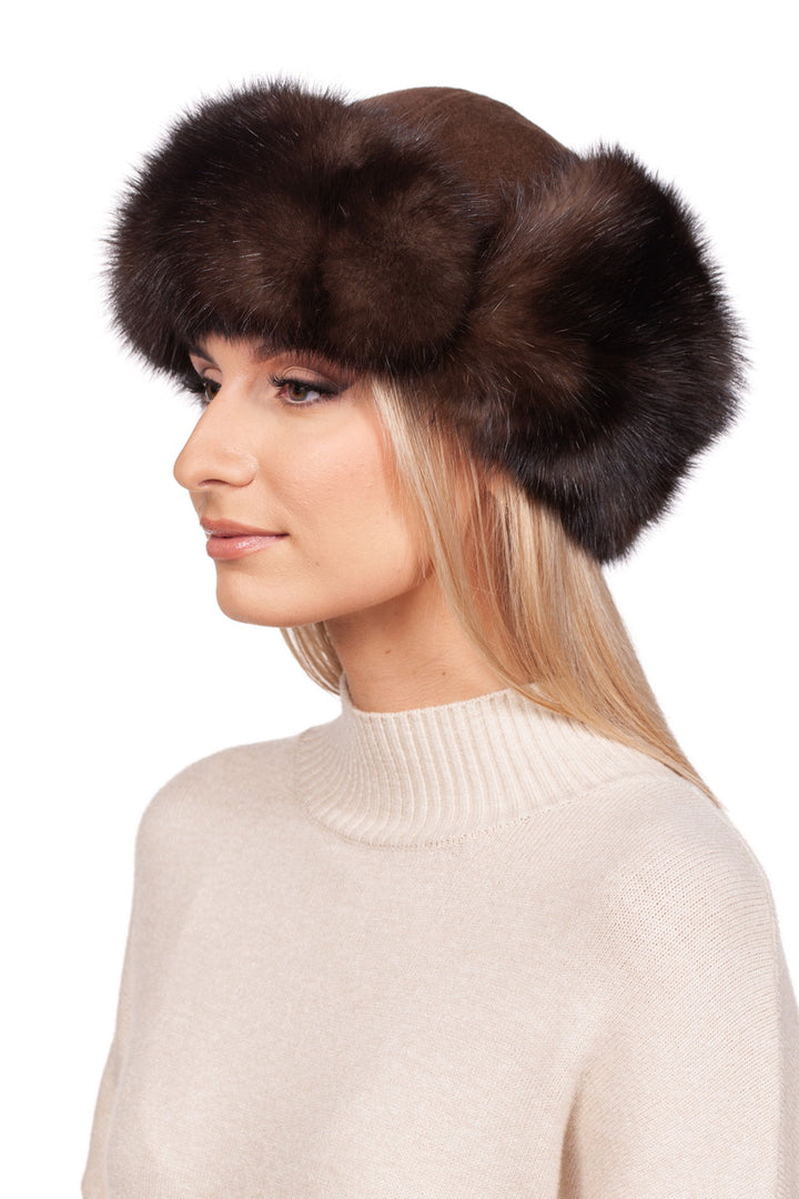 A woman with long blonde hair is wearing a luxurious winter accessory, the Brown Barguzin Sable Fur Ushanka Hat by FurbySD, along with a light cream turtleneck sweater. The background is plain white. Her expression is neutral as she gazes to the side.