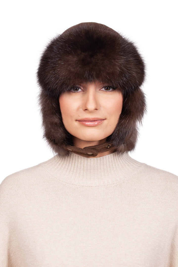 Dressed in a cozy light-colored sweater, a person smiles at the camera while wearing a luxurious Brown Barguzin Sable Fur Ushanka Hat by FurbySD. The large, fluffy winter accessory elegantly covers their ears against the plain white background.