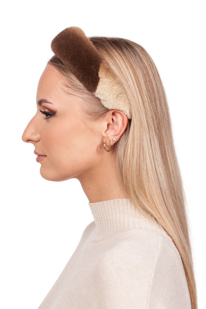 A woman with long blonde hair is elegantly wearing the Brown Sheared Mink Fur Hairband by FurbySD, a luxurious handmade accessory in a fluffy, two-toned brown design. She's paired it with a cream sweater and has multiple earrings in her left ear, all set against a plain white background while facing to the left.