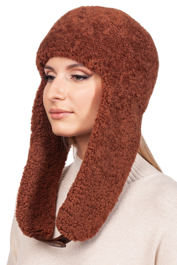 A person wearing the FurbySD Rusty Brown Shearling Fur Earflap Hat gazes to the left. Their long hair cascades over a light-colored sweater, creating a contrast against the plain white background. This cozy winter accessory adds warmth and style to their ensemble.