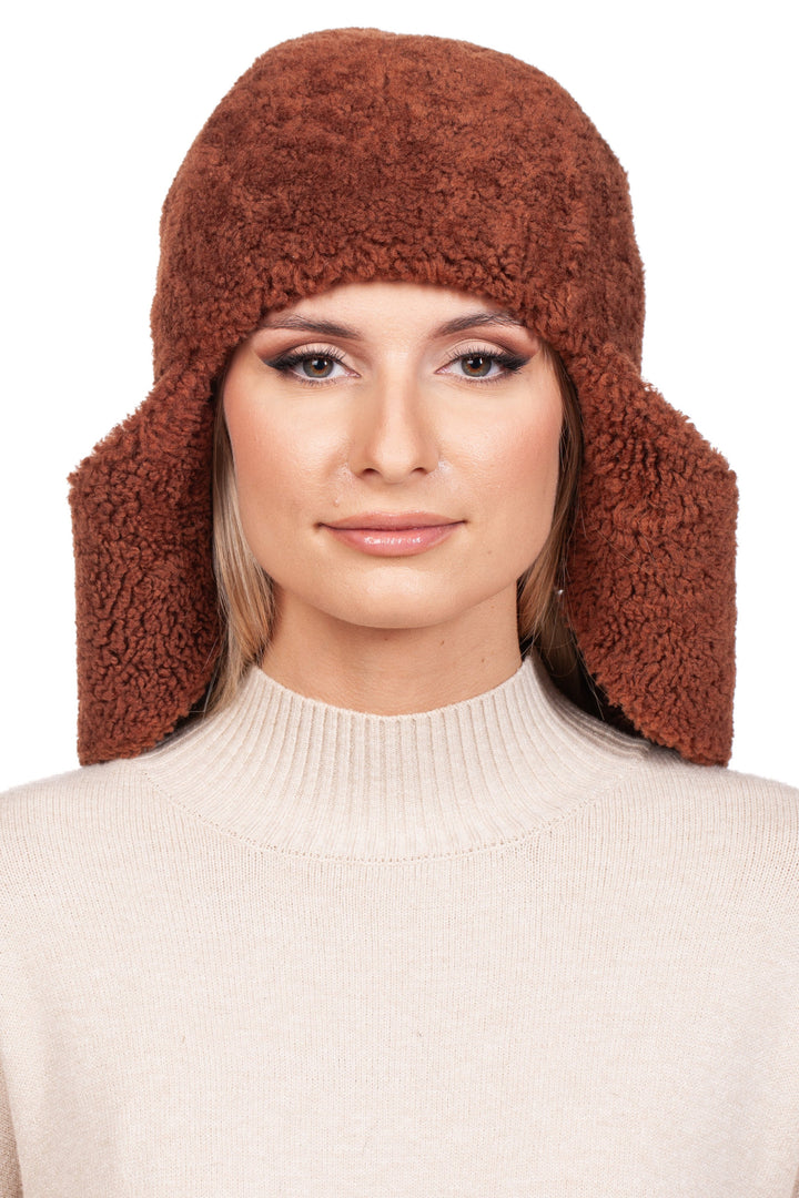 A woman wearing the FurbySD Rusty Brown Shearling Fur Earflap Hat over a light beige turtleneck sweater looks directly at the camera with a neutral expression. The plain white background accentuates this chic winter accessory.