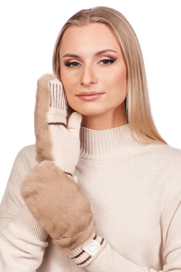 A woman with long blonde hair is wearing a cream-colored turtleneck and soft beige flip top mittens with faux fur cuffs from FurbySD. Complementing her outfit, a beige mink fur hat from the Beige Mink Fur Hat And Flip Top Mittens Set sits elegantly atop her head. She touches her cheek with one hand, looking directly at the camera with a neutral expression.