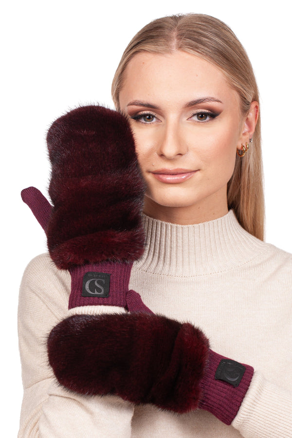 A person with long hair wears a cream-colored sweater and small hoop earrings, holding one mitten to their face with a calm expression. They are wearing Burgundy Red Mink Fur Flip Top Mittens from FurbySD, which feature a logo patch on the cuff.