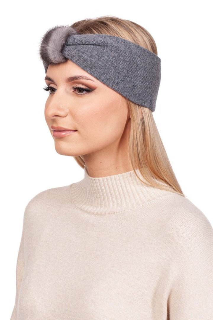 A woman with long blonde hair is wearing the FurbySD Grey Cashmere Headband with Mink Fur, which features a mink fur accent knot at the front. She is dressed in a light beige turtleneck sweater and gazes to the side against a plain white background.