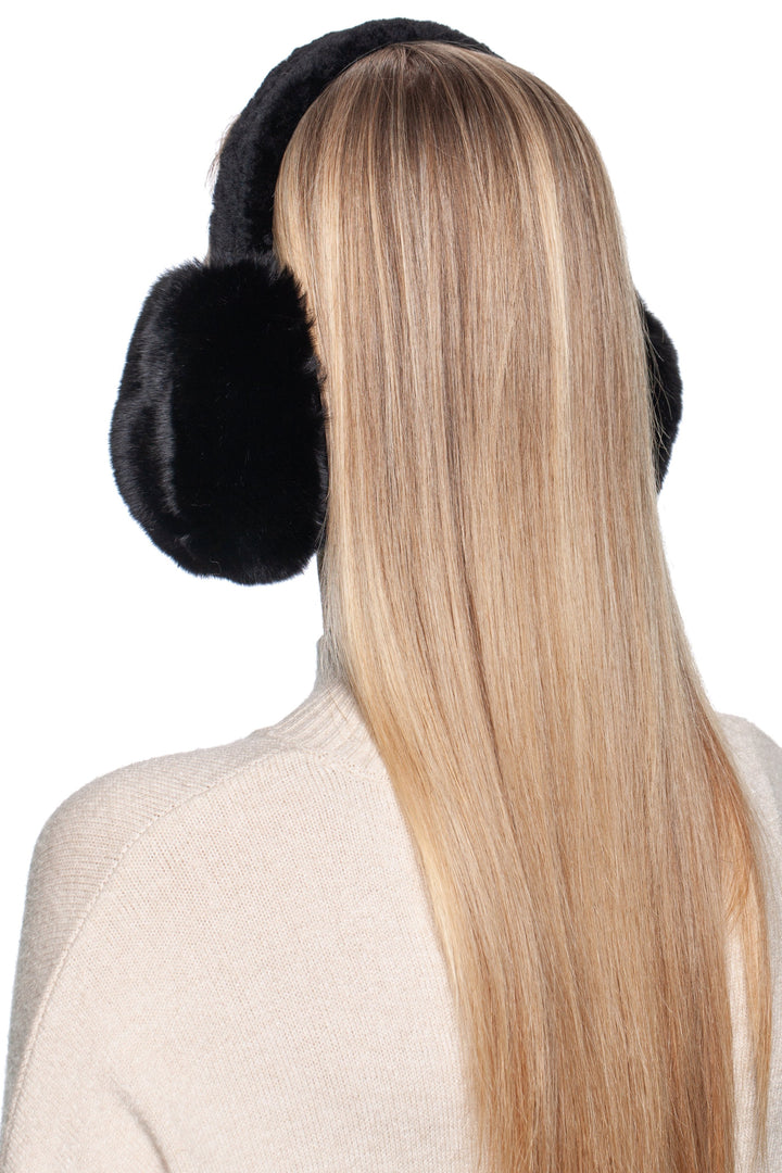 A person with long, straight blonde hair is wearing FurbySD's Black Chinchilla Fur Earmuffs with Astrakhan Band. They are standing against a plain white background, facing away from the camera, in a cream-colored sweater.