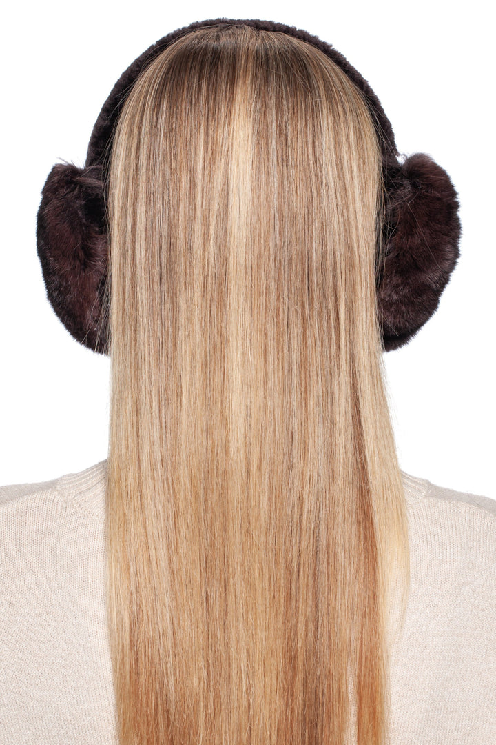 A person with long blonde hair is seen from the back, donning luxurious FurbySD Brown Chinchilla Fur Earmuffs With Astrakhan Fur and a light-colored sweater. These earmuffs serve as an elegant winter accessory against the pristine white backdrop.