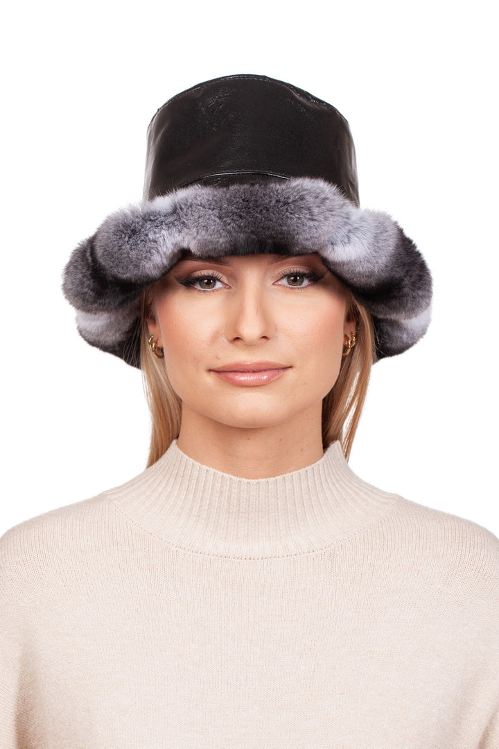 A person is wearing a Chinchilla Fur Panama Hat by FurbySD, paired with a beige turtleneck sweater. This luxury accessory, featuring Nappa leather and gray fur trim, frames their softly smiling face against a plain white background.