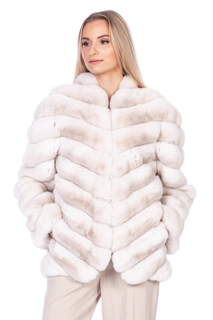 A person with long hair is wearing a FurbySD Cream Chinchilla Fur Coat and beige trousers. They stand with a neutral expression, hands in pockets, and wearing high heels, exuding an aura of elegance.